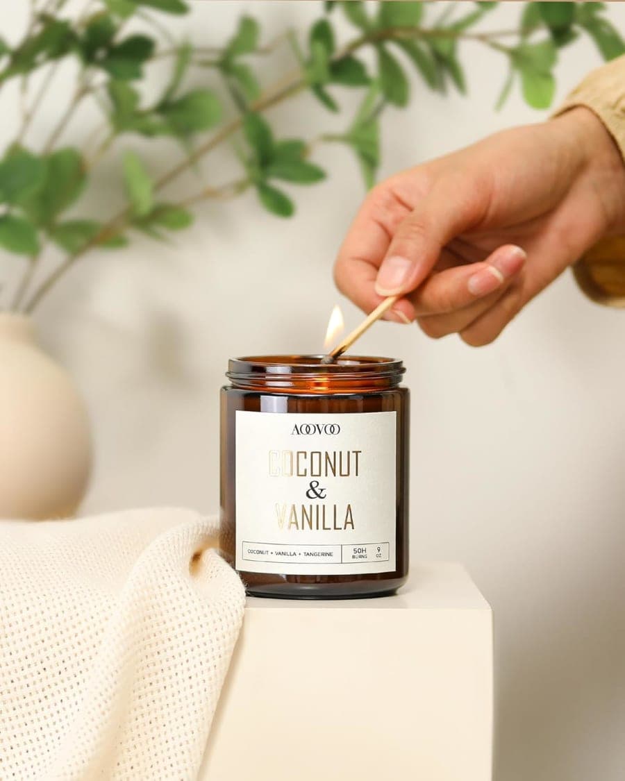 Amber jar coconut and vanilla candle being lit beside a soft cream towel and leafy greenery.