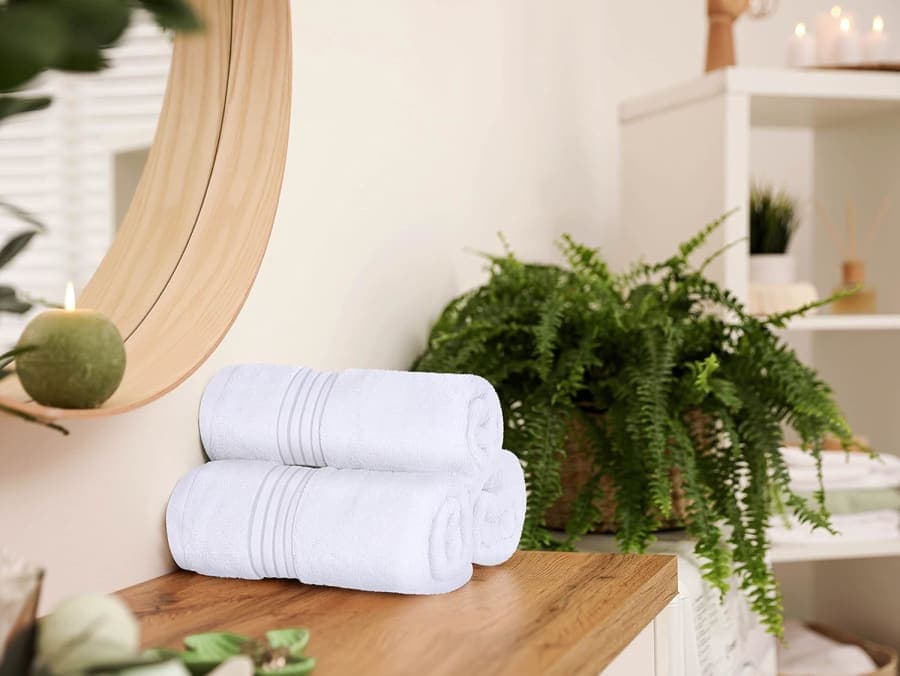 Three rolled white cotton hand towels displayed on a wooden bathroom vanity beside a round mirror and green fern.